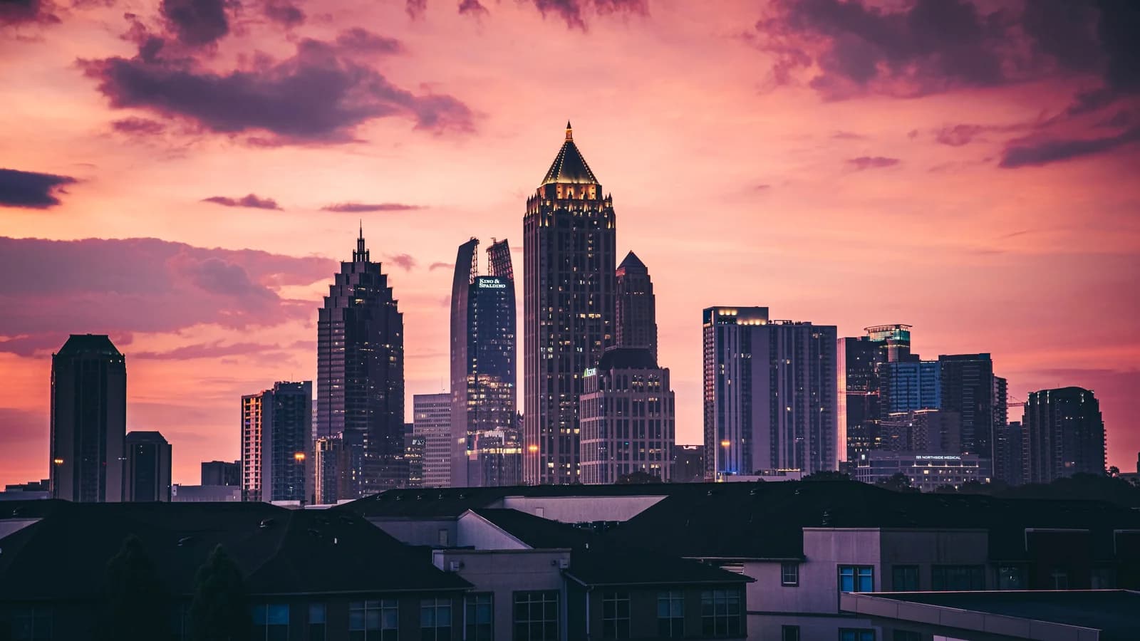 Midtown Atlanta skyscrapers, led by a pointed lit tower, silhouetted against a pink and orange sunset above rooftops