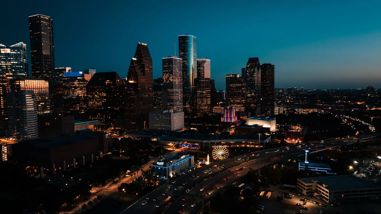 Downtown Houston skyscrapers lit up at dusk under a deep blue sky, with a glowing Ferris wheel and a busy freeway below