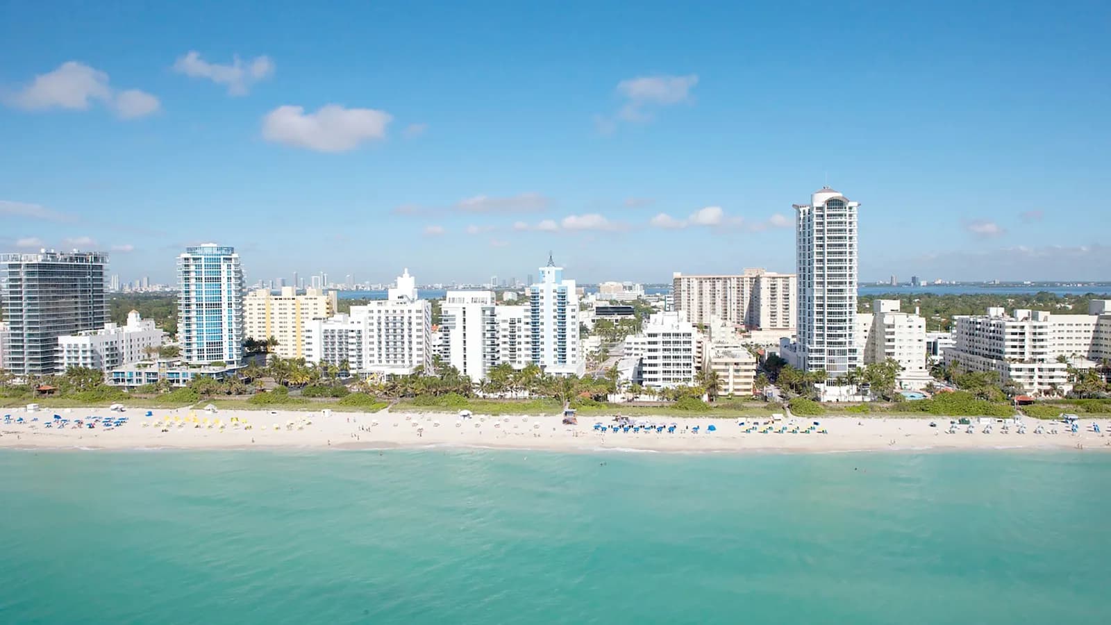 Aerial view of Miami Beach, with white high-rise towers and palms behind a wide strip of sand dotted with umbrellas, turquoise ocean in front and a blue sky above