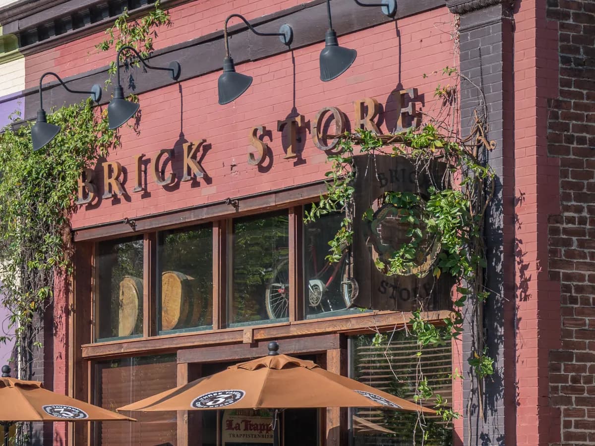 The red brick front of a pub with Brick Store in raised letters, black gooseneck lamps, climbing vines and tall windows above patio umbrellas