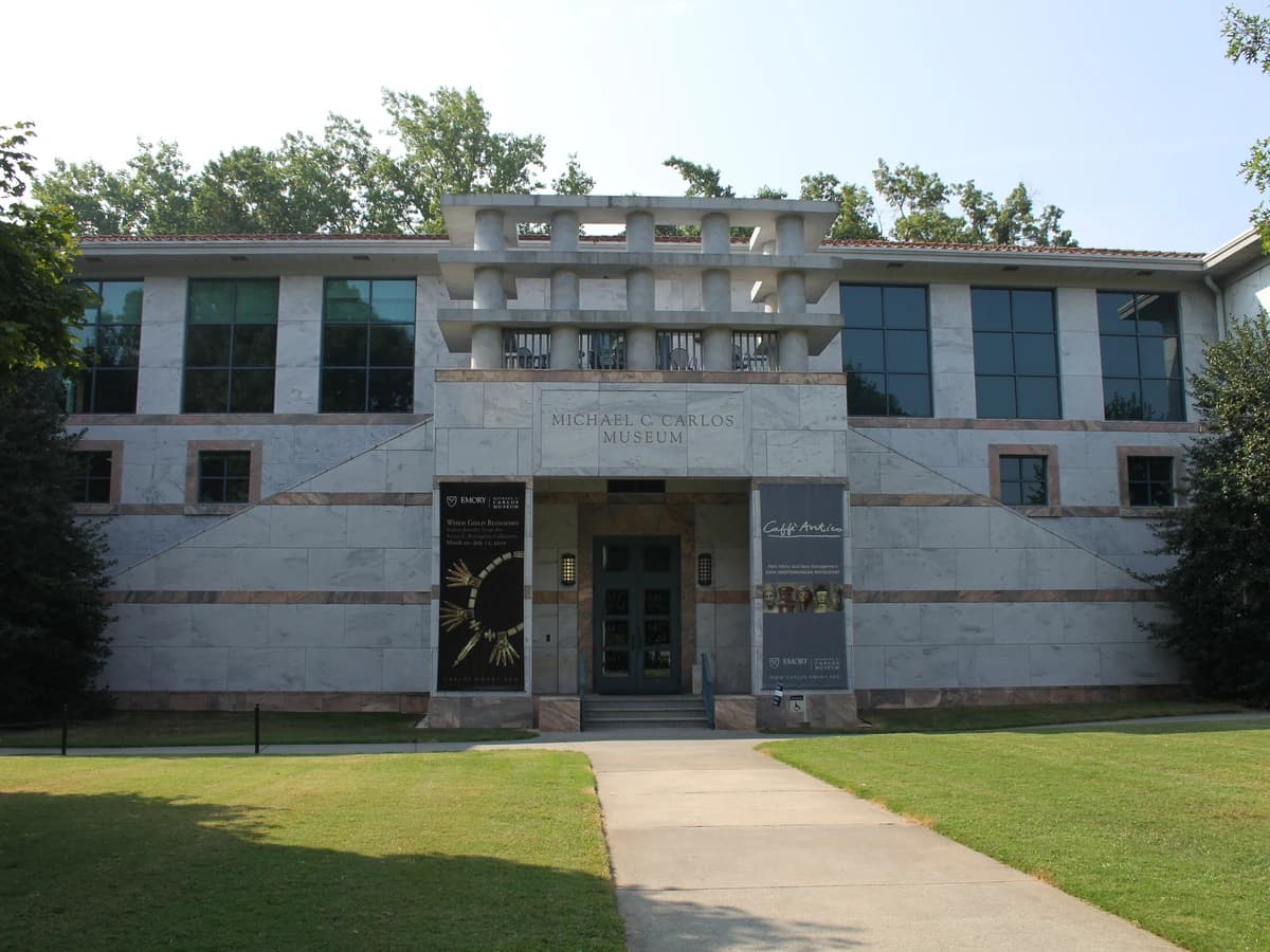 A pale building banded in gray and white marble with a central entrance, behind a green lawn and a straight paved walk under trees