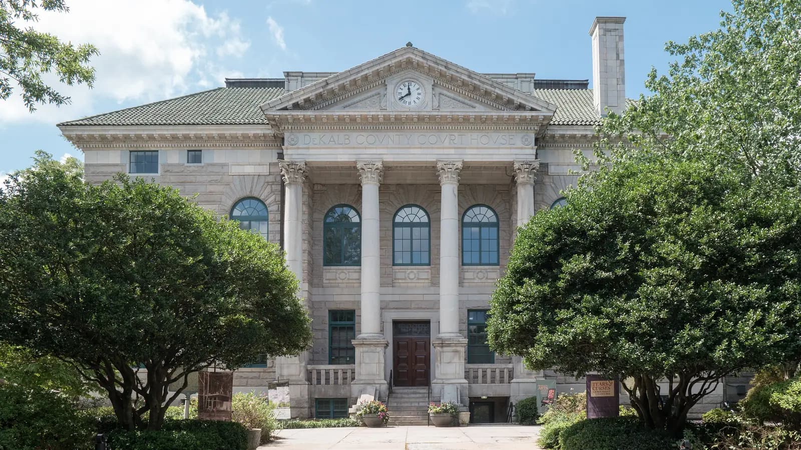 The granite Historic DeKalb Courthouse in Decatur, with a columned portico, a clock in the pediment and leafy trees framing the front steps