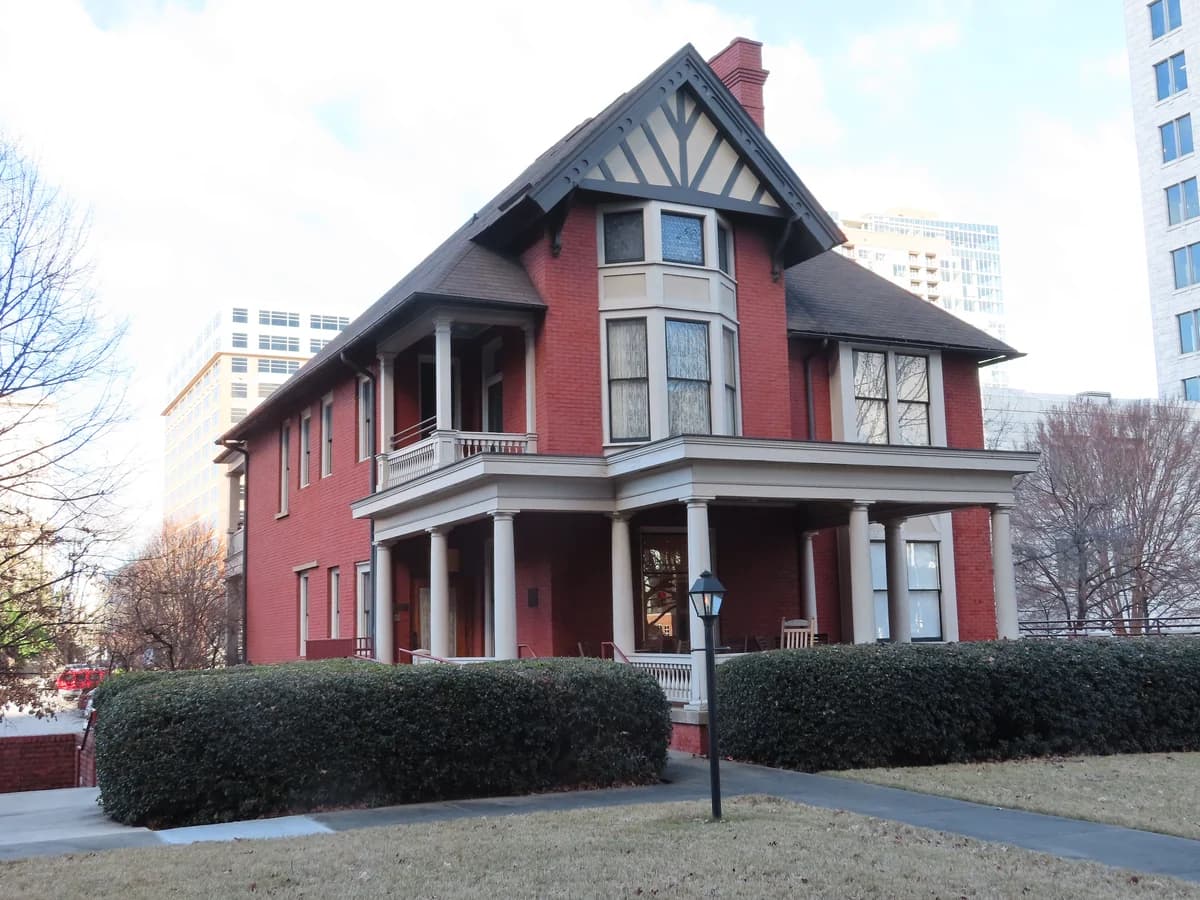 A red brick and timber Tudor style house with bay windows and a white columned porch behind trimmed hedges, with taller buildings behind