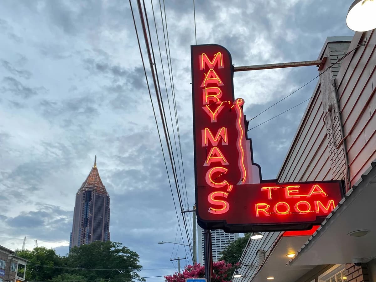 Red neon sign reading Mary Mac's Tea Room on a bracket above a brick building's awning, with a tall pointed office tower under a cloudy sky