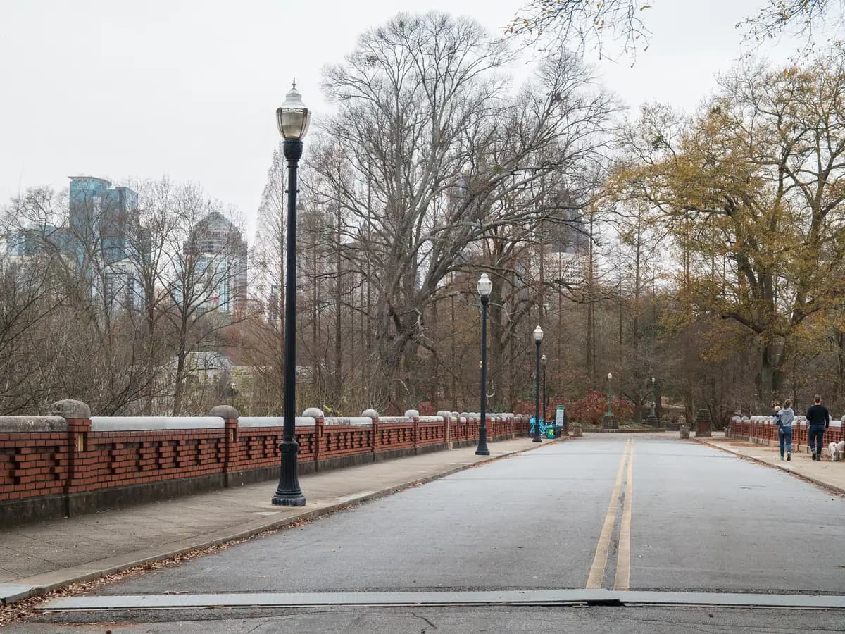 A road across a brick-walled bridge with old lampposts leading into a park of bare winter trees, with city towers rising behind