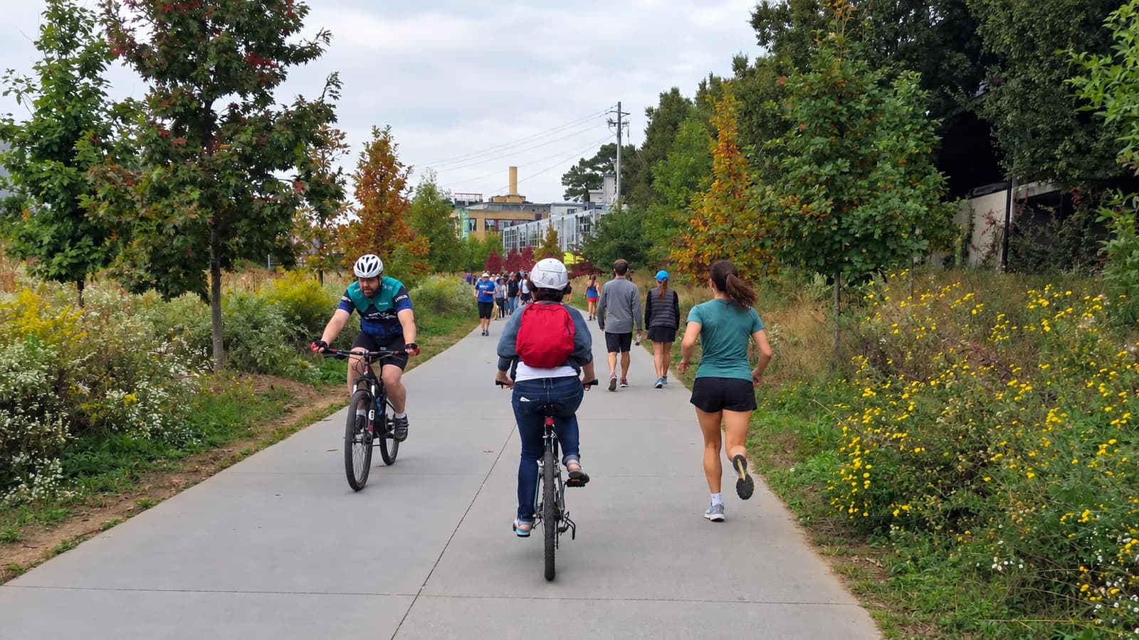 A runner in a teal shirt on the BeltLine Eastside Trail beside cyclists, with yellow wildflowers and young trees lining the path