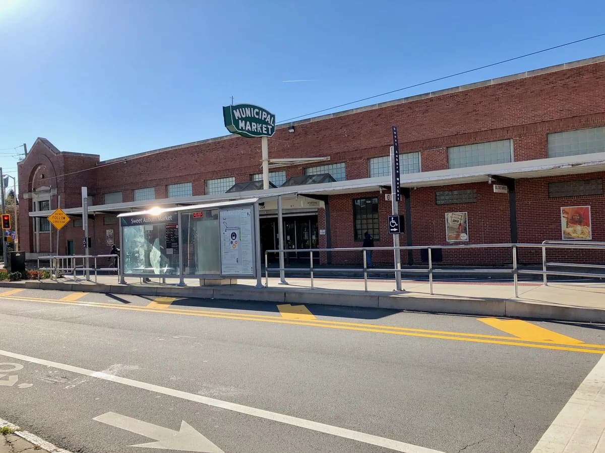 A long red brick market hall with a green Municipal Market sign and a covered streetcar stop in front, seen across an empty street on a sunny day