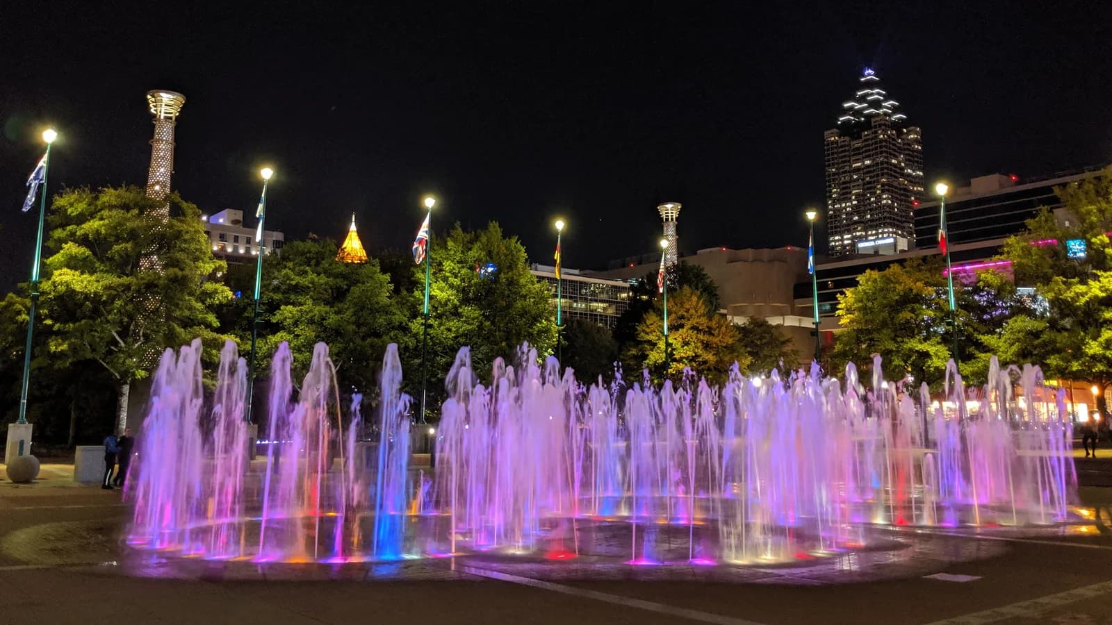 Jets of water lit pink and purple rise from the Fountain of Rings in Centennial Olympic Park at night, with trees, flag poles and a lit downtown tower behind