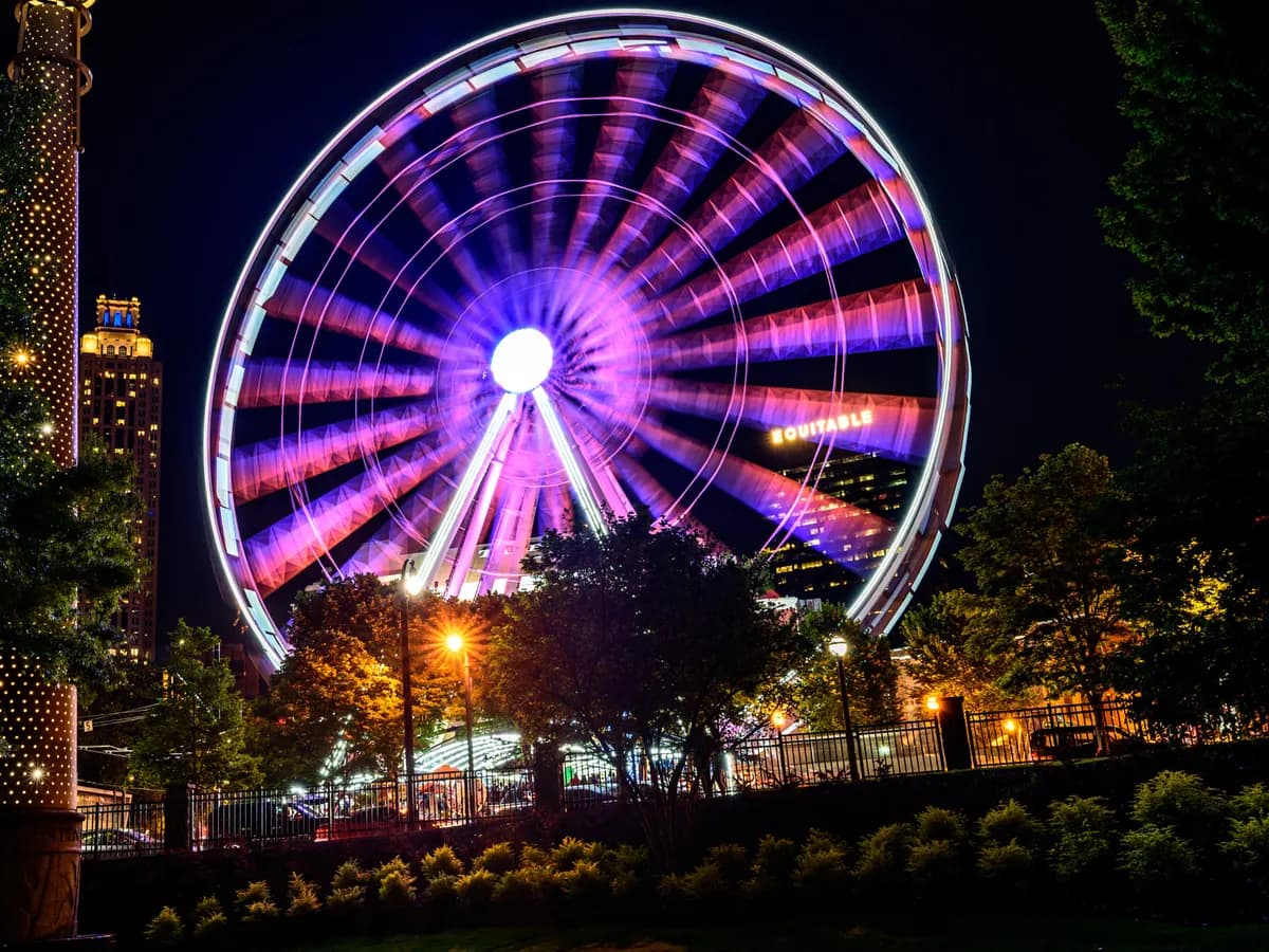 A large observation wheel glowing purple and pink against the night sky, with lit trees, a fence and an office tower behind it