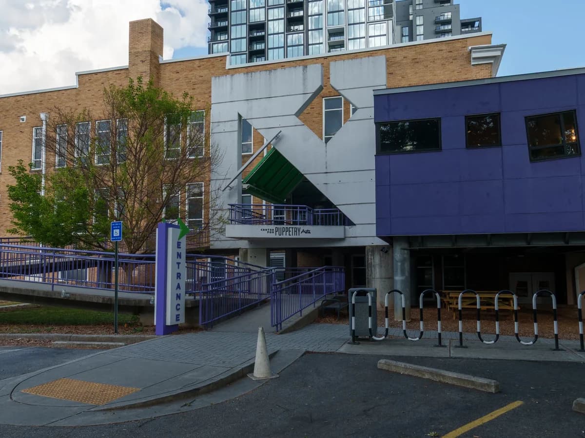 A purple-clad modern wing and a white entrance block with angled panels beside an older tan brick building, with a ramp and driveway in front
