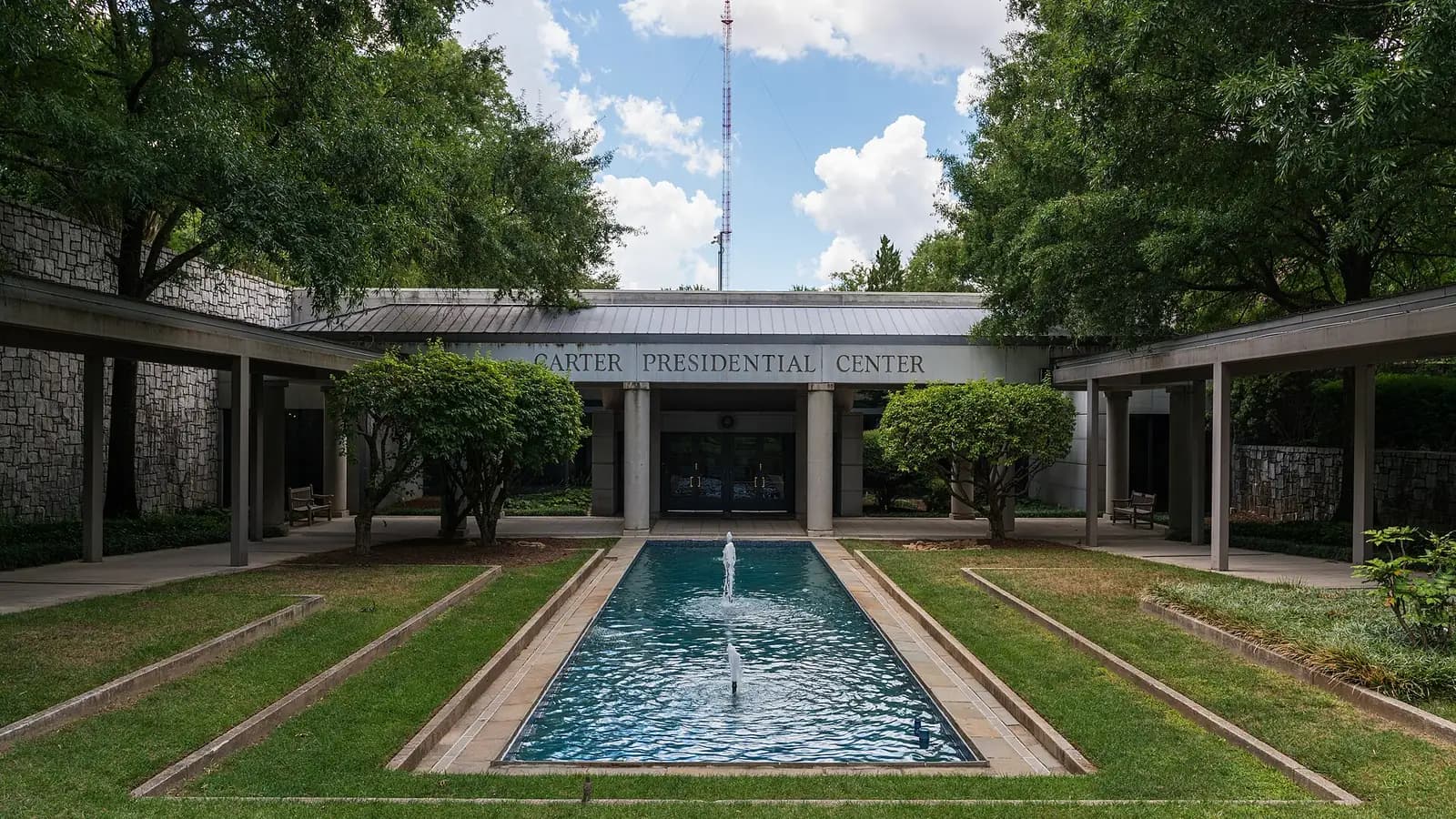 A long reflecting pool with a fountain between lawns and covered walkways, leading to the entrance of the Carter Presidential Center in Atlanta, framed by trees