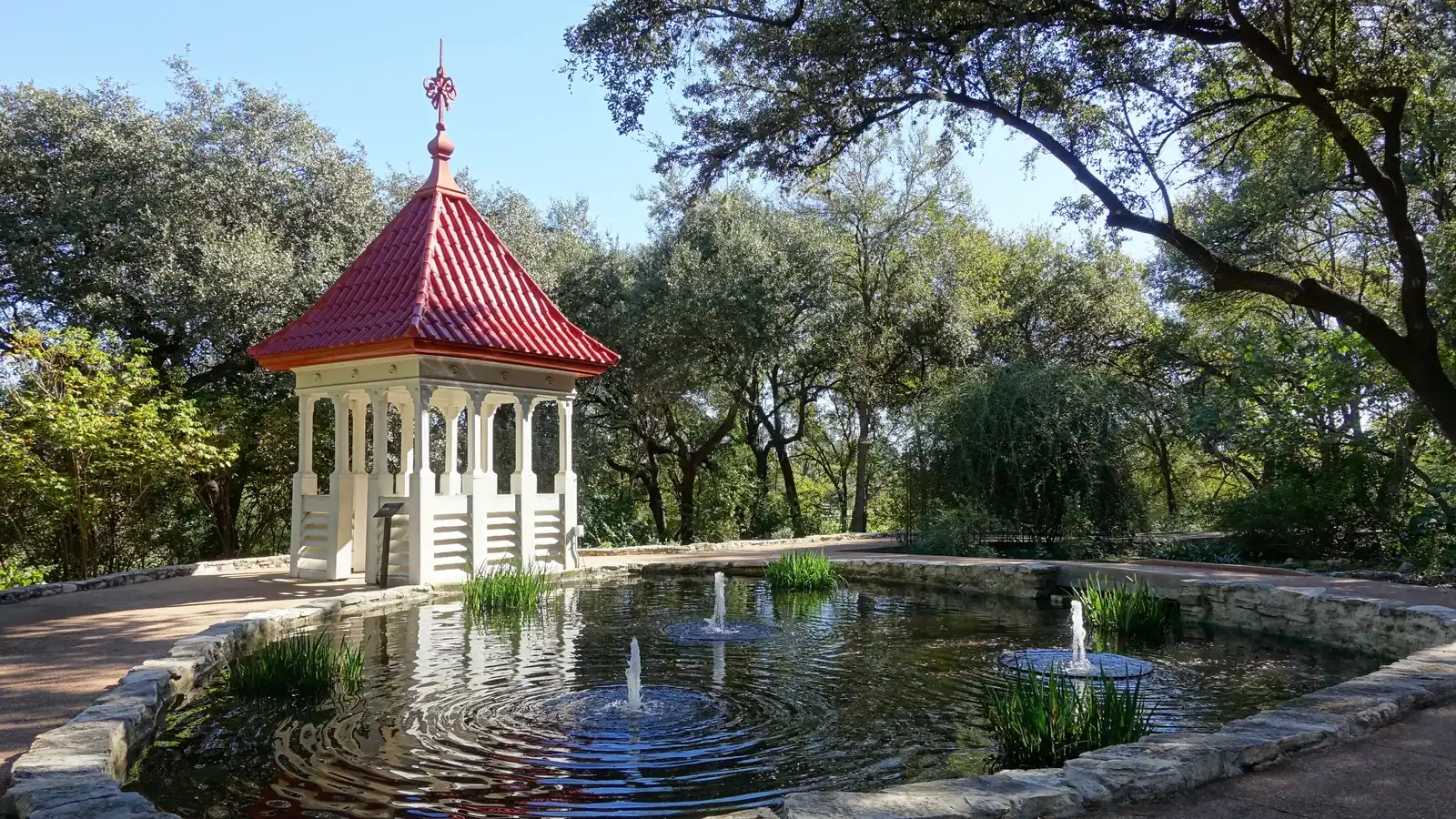 A white gazebo with a red tiled roof beside a round stone-edged pond with small fountains, under live oaks at Zilker Botanical Garden
