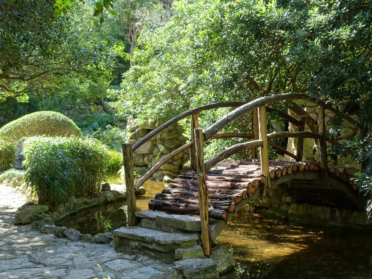 A rustic wooden footbridge with log railings arches over a still stream in a shady Japanese garden, beside stone steps, a flagstone path and leafy trees