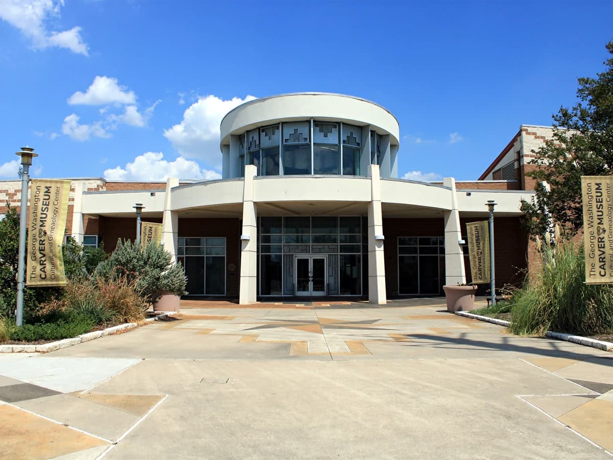 A modern museum entrance with a round glass upper floor over white columns and glass doors, beside banners and plantings on a sunny day