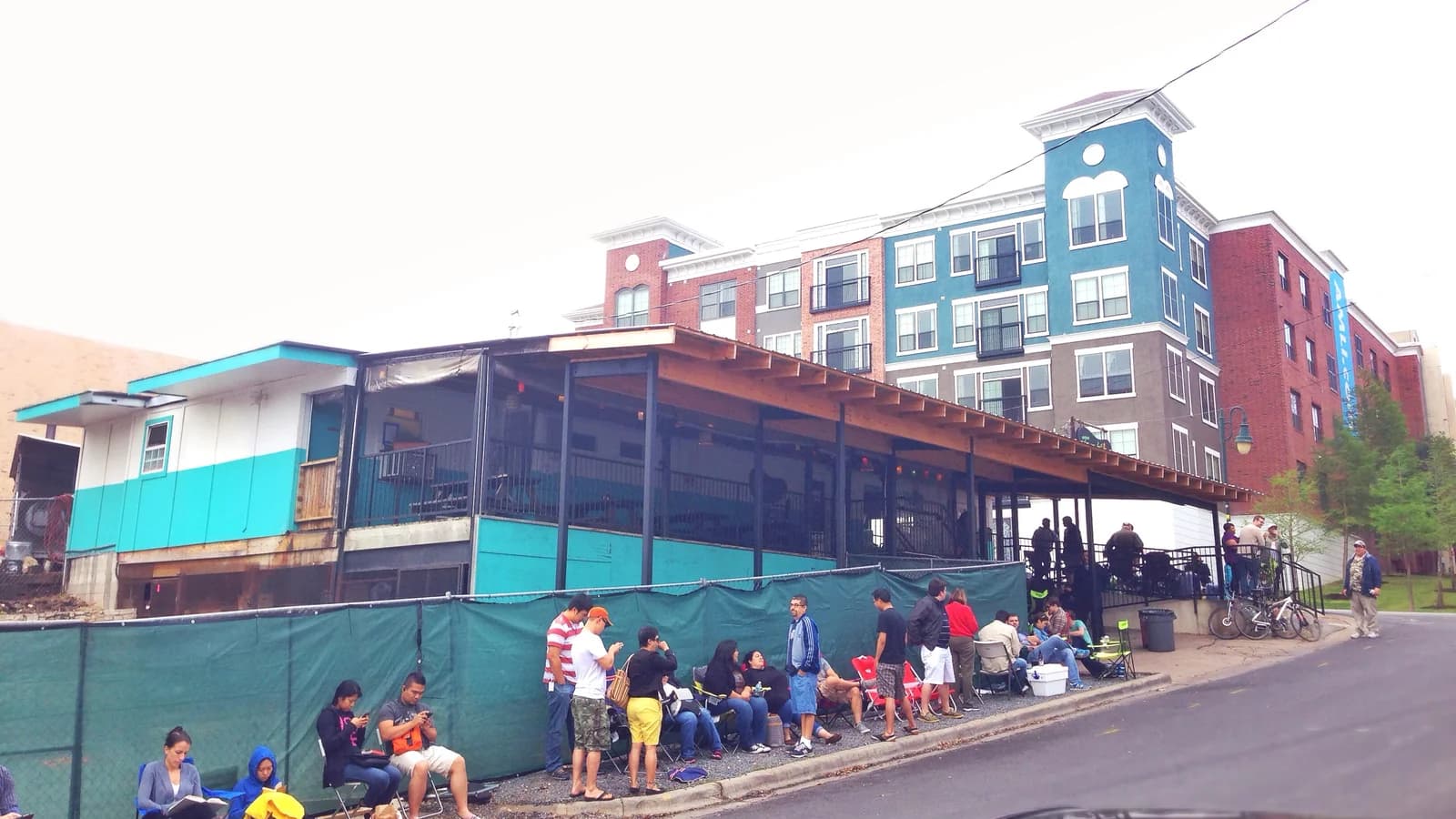 A line of people with folding chairs waiting beside a green fence outside the low teal Franklin Barbecue building on an overcast morning, with apartments behind