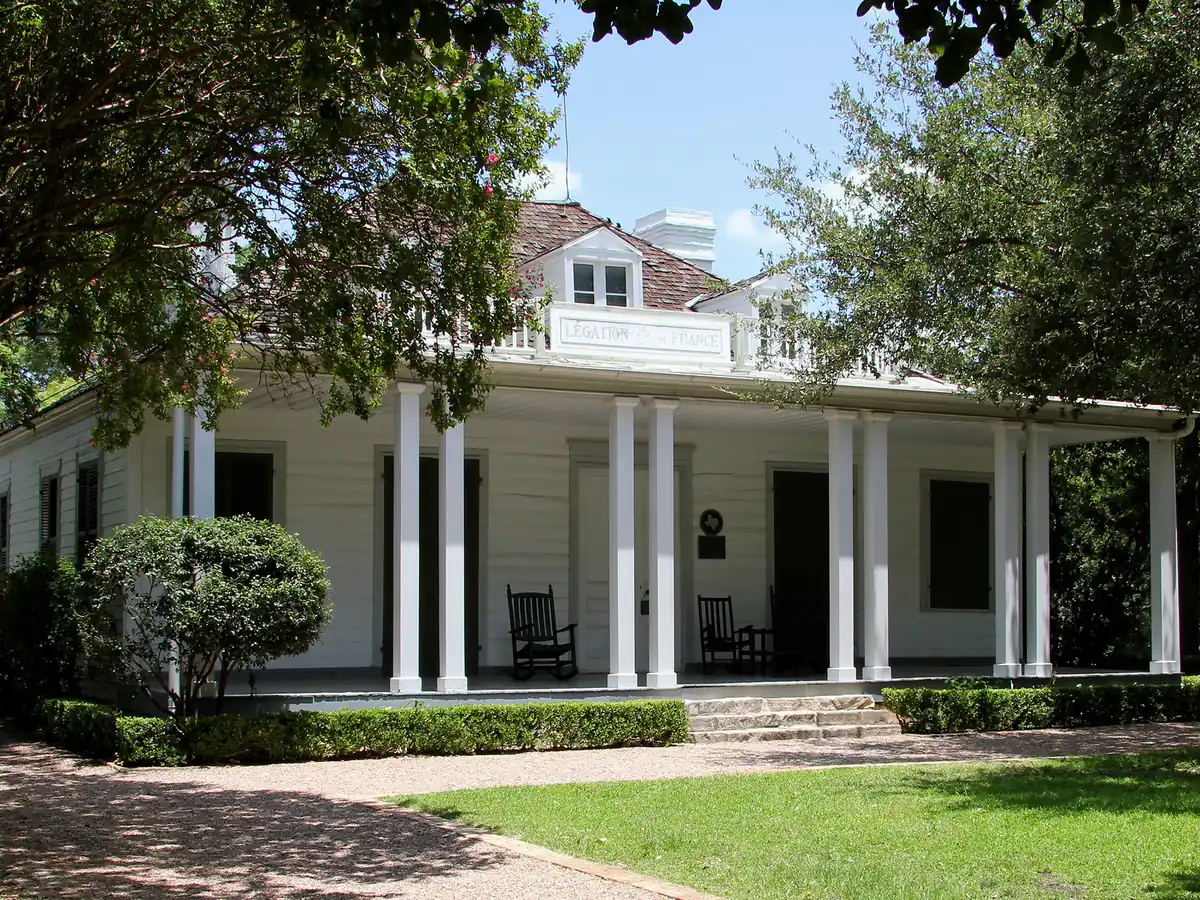 A white wooden house with a long porch of white columns, rocking chairs and a sign on the roof balcony, behind a trimmed hedge, a gravel path and a lawn