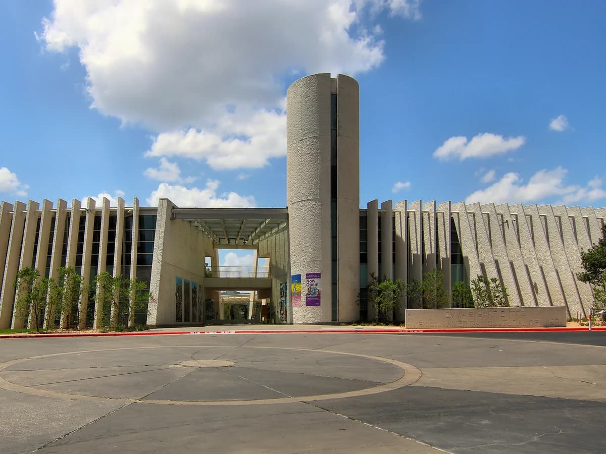 A wide pale building with a ribbed facade and a tall round tower over a glass entrance, behind an open paved plaza under a blue sky