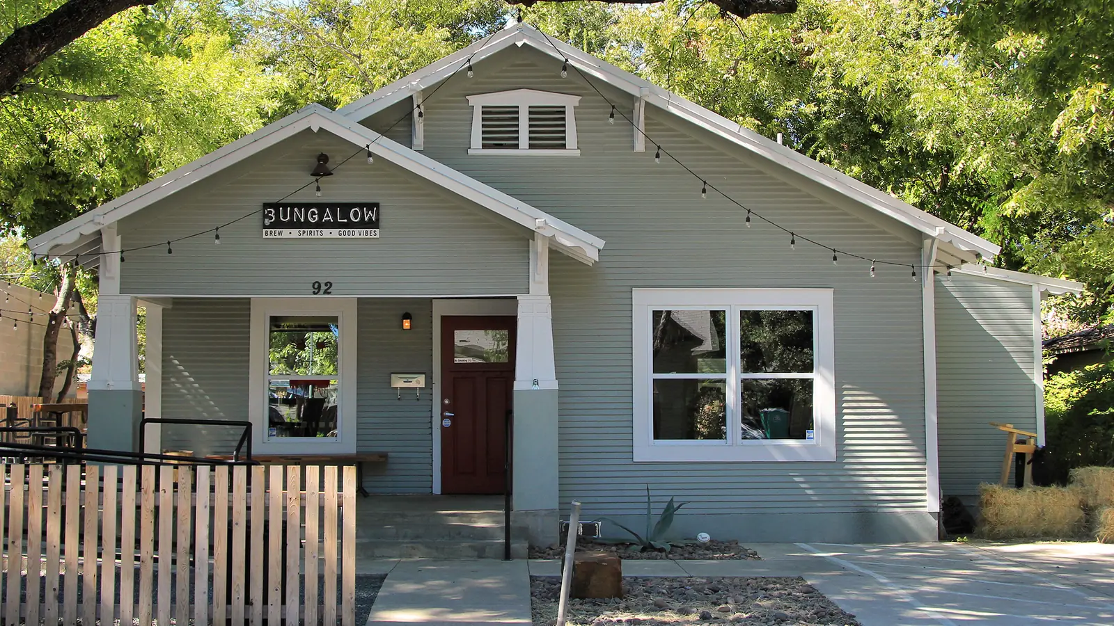 A gray wooden bungalow with a deep front porch and string lights along its eaves, now a bar called Bungalow, shaded by trees on Rainey Street