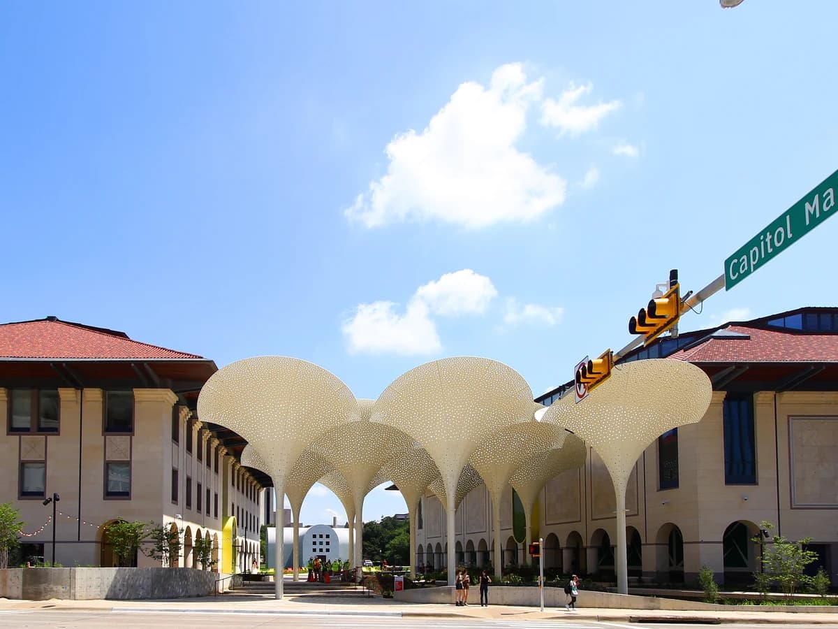 A row of tall white petal shaped canopies on a plaza between two pale buildings with red tile roofs, beside a street sign on a sunny day