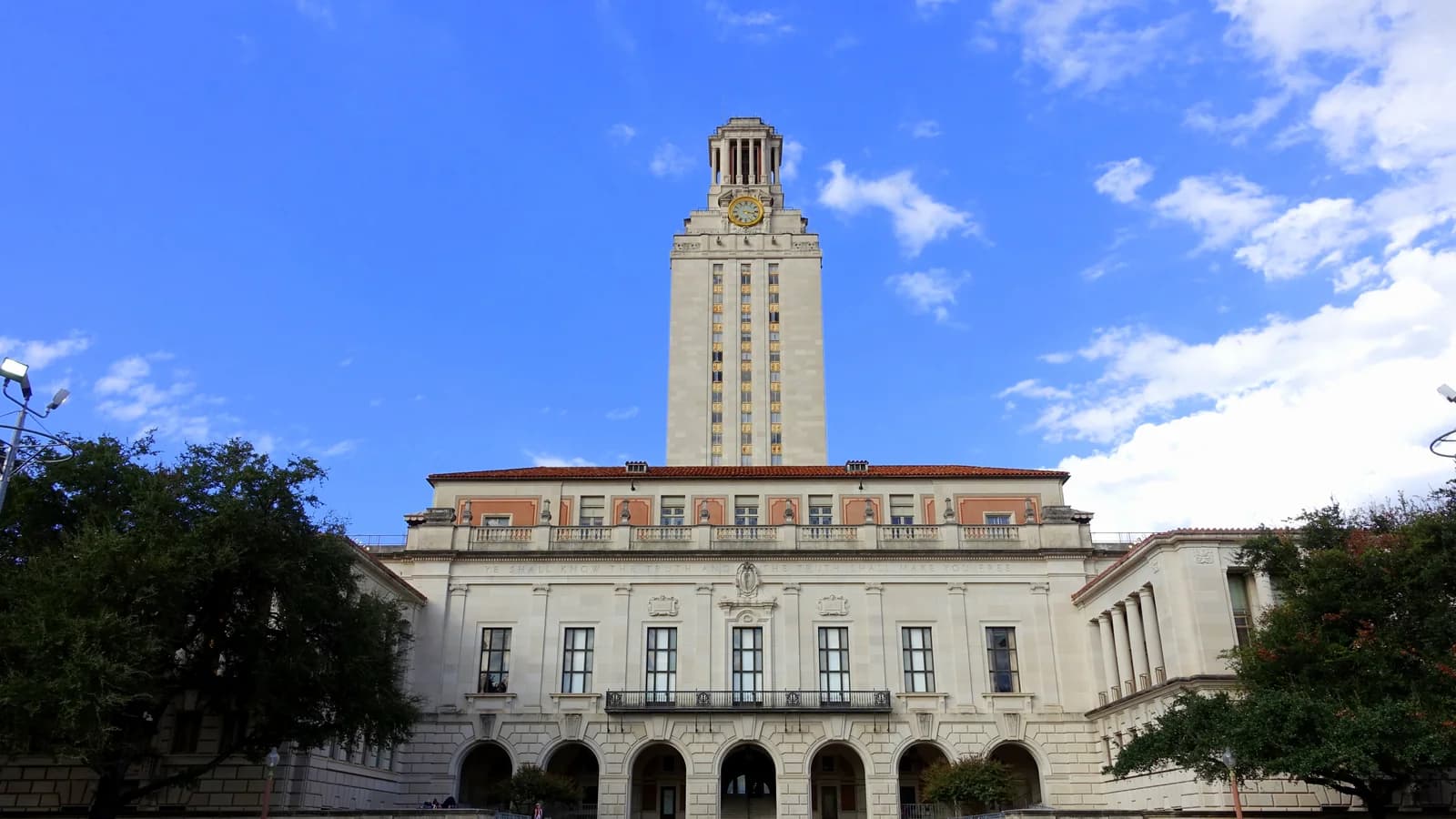 The UT Tower rising above the columned Main Building of the University of Texas at Austin, with oak trees under a blue sky