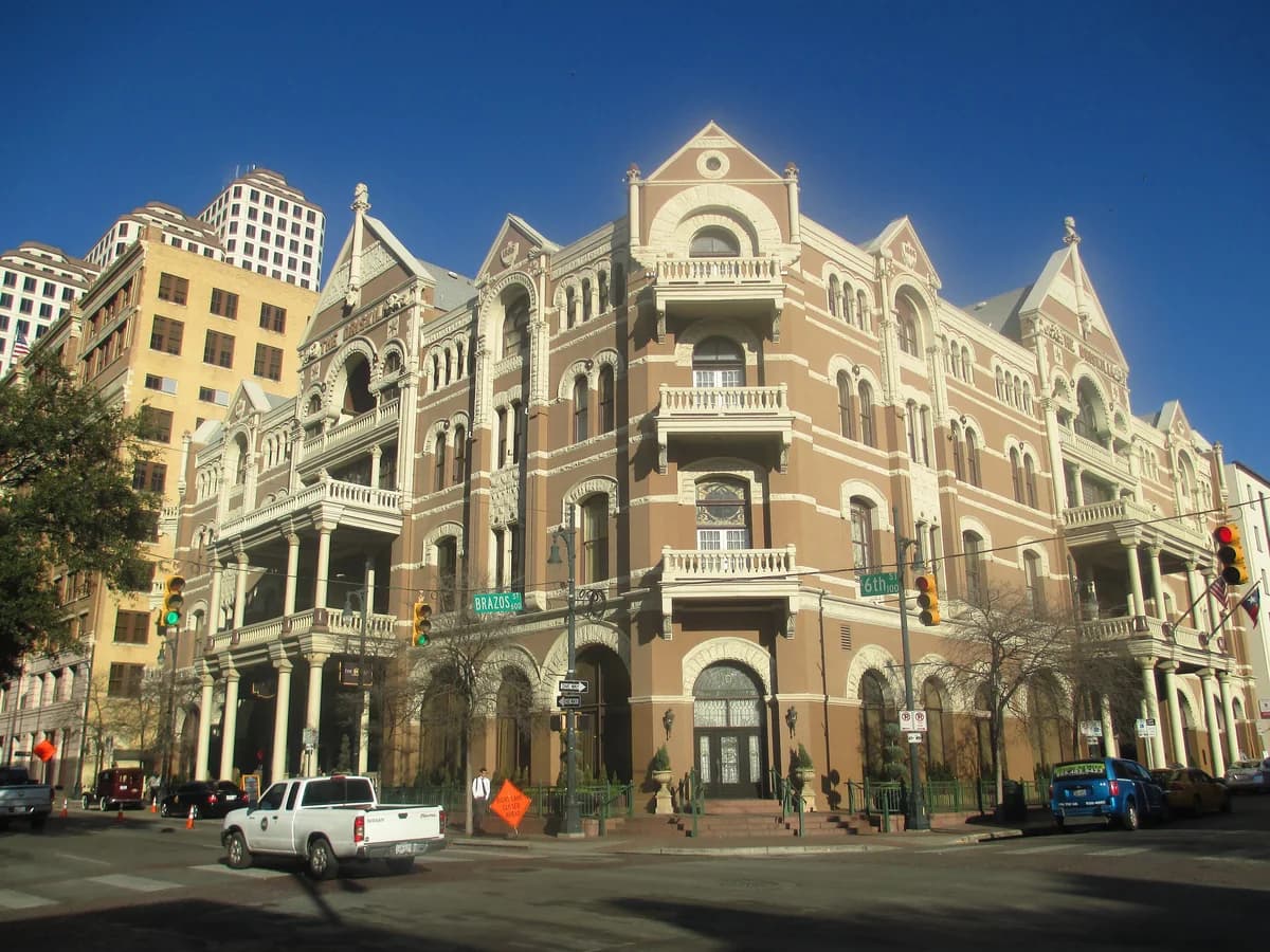 An ornate four story tan brick hotel with arched balconies and rooftop towers on a street corner under a deep blue sky, with a white pickup in front