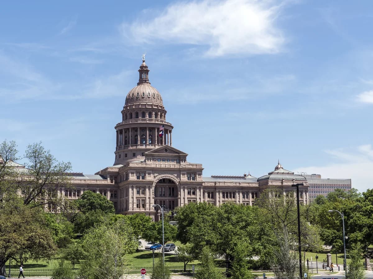 A domed granite capitol building rising above green trees and lawns on a sunny day, with lampposts lining a path in front