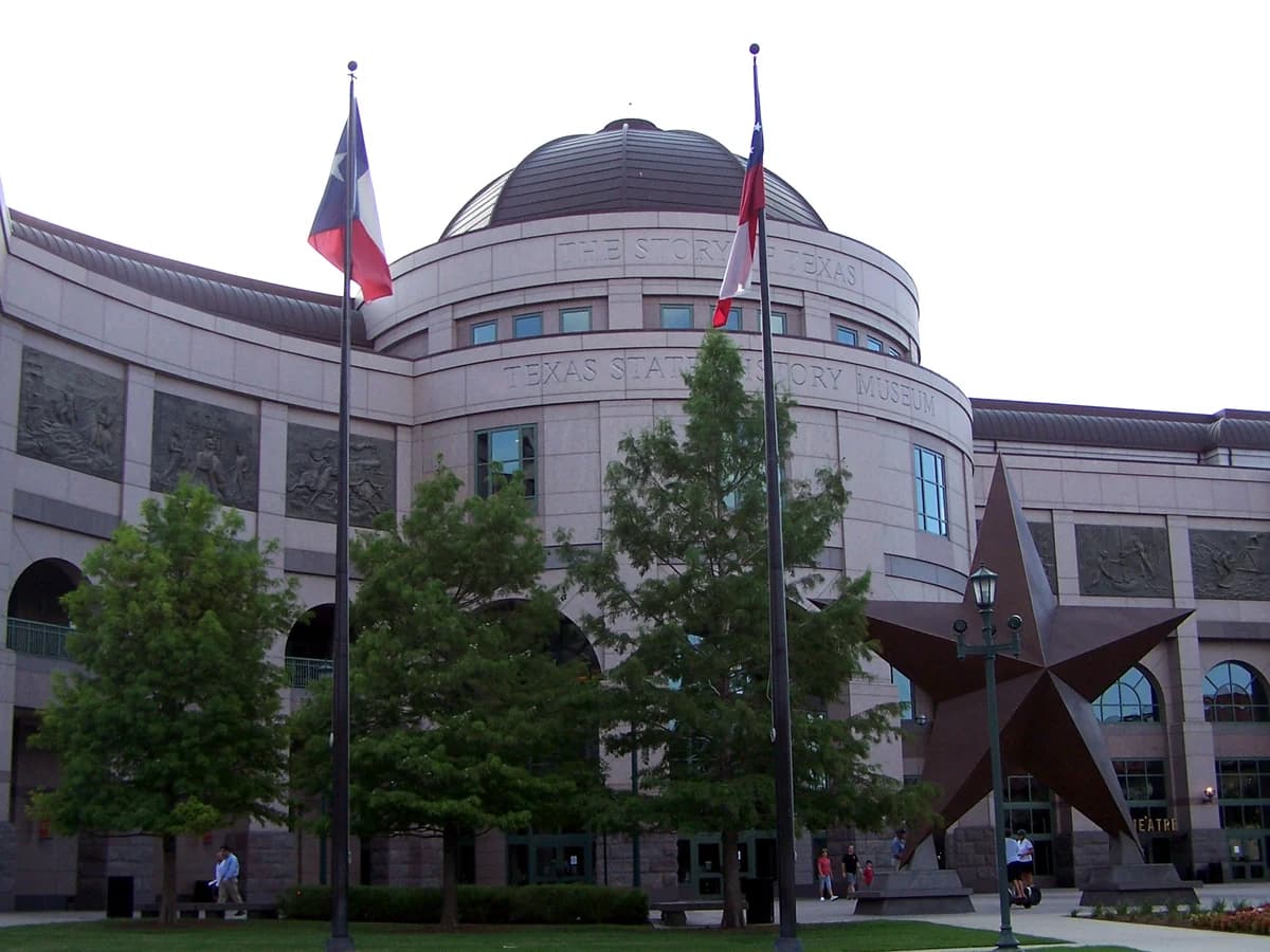 A domed stone museum with a curved front, two flagpoles and a giant bronze star sculpture on the lawn under a pale sky