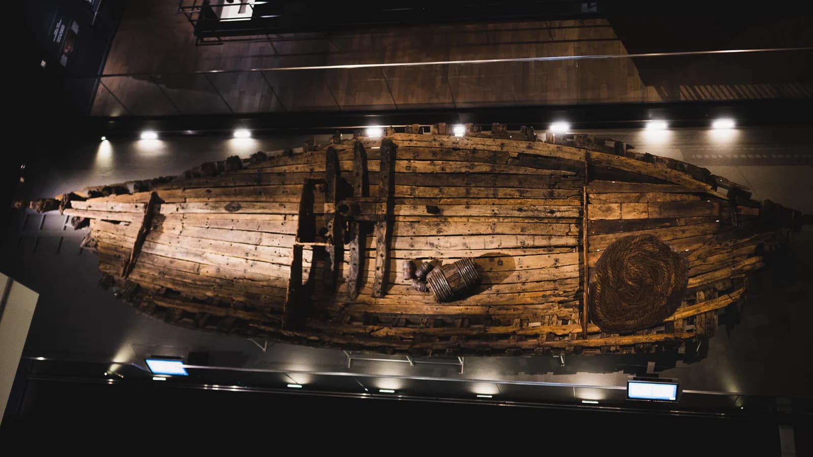 The reassembled wooden hull of the 17th century French ship La Belle seen from above, lit by a row of spotlights in a dim museum gallery