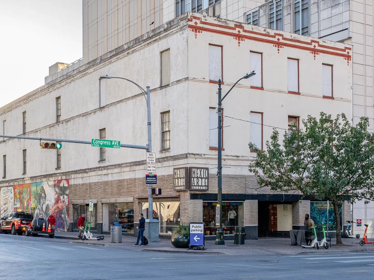 A pale three story building with a red trimmed roofline and a colorful mural at street level on a downtown corner, with towers rising behind