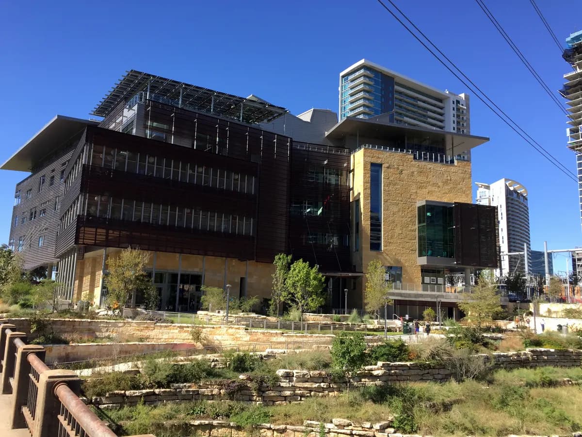 A modern building of glass, metal screens and stone above a rocky garden of native plants, with tall towers in the background