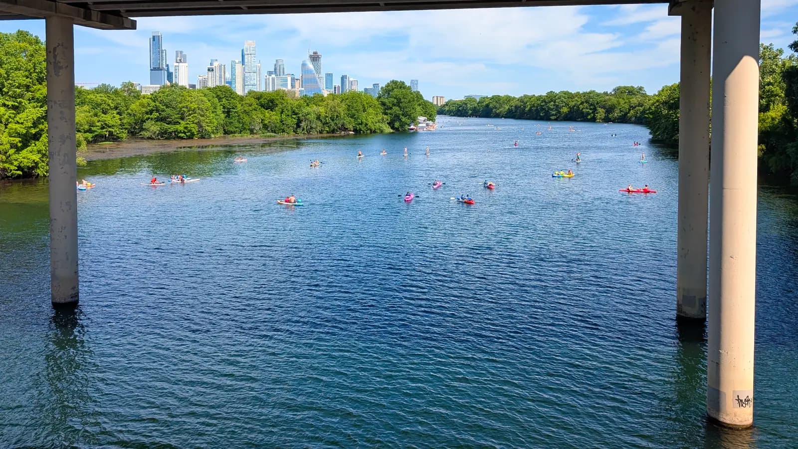 Kayakers and paddleboarders spread across blue water on Lady Bird Lake, framed by bridge pillars, with downtown towers rising behind the trees