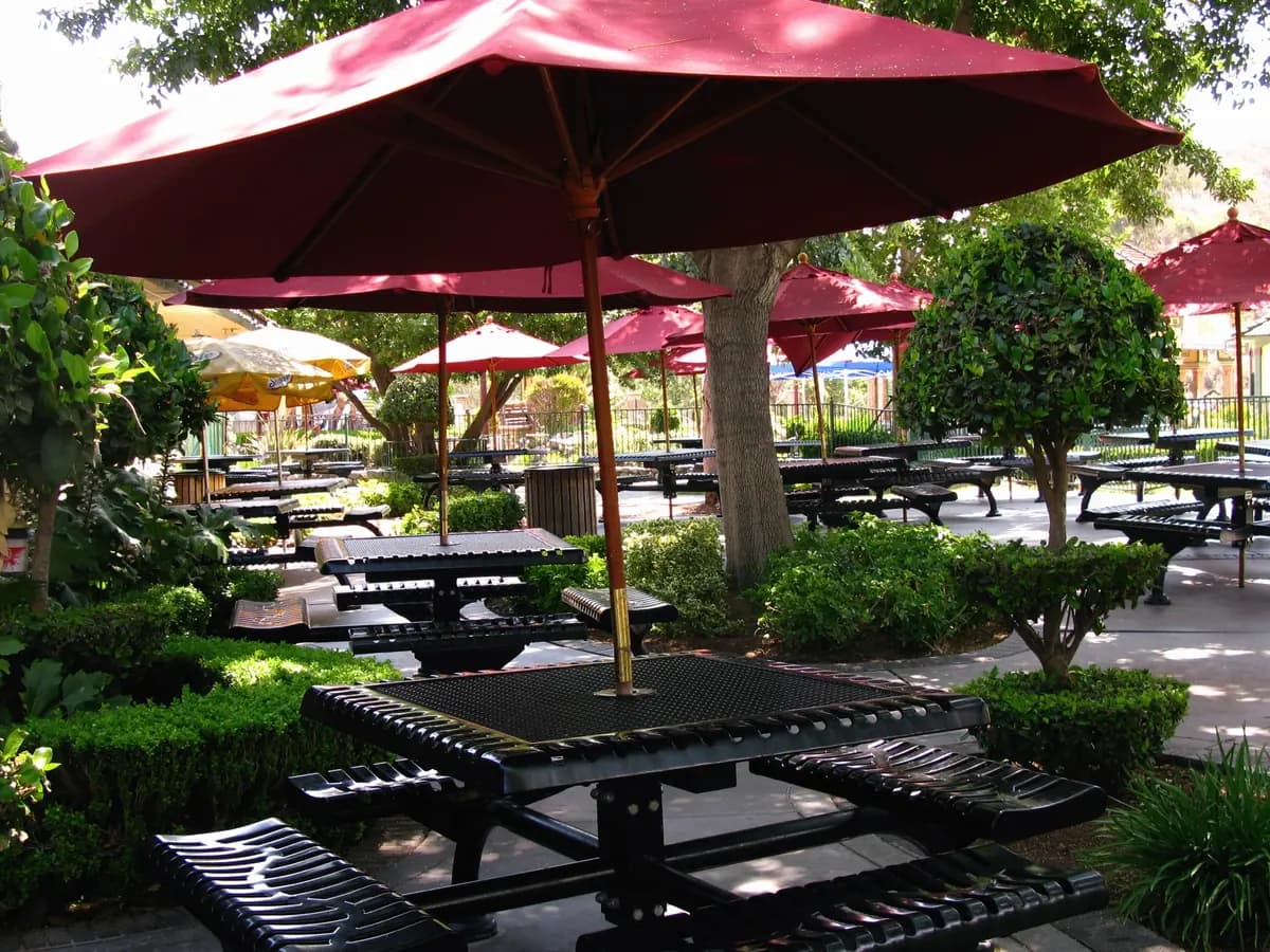Rows of dark picnic tables under red patio umbrellas in a leafy outdoor beer garden, with no one seated