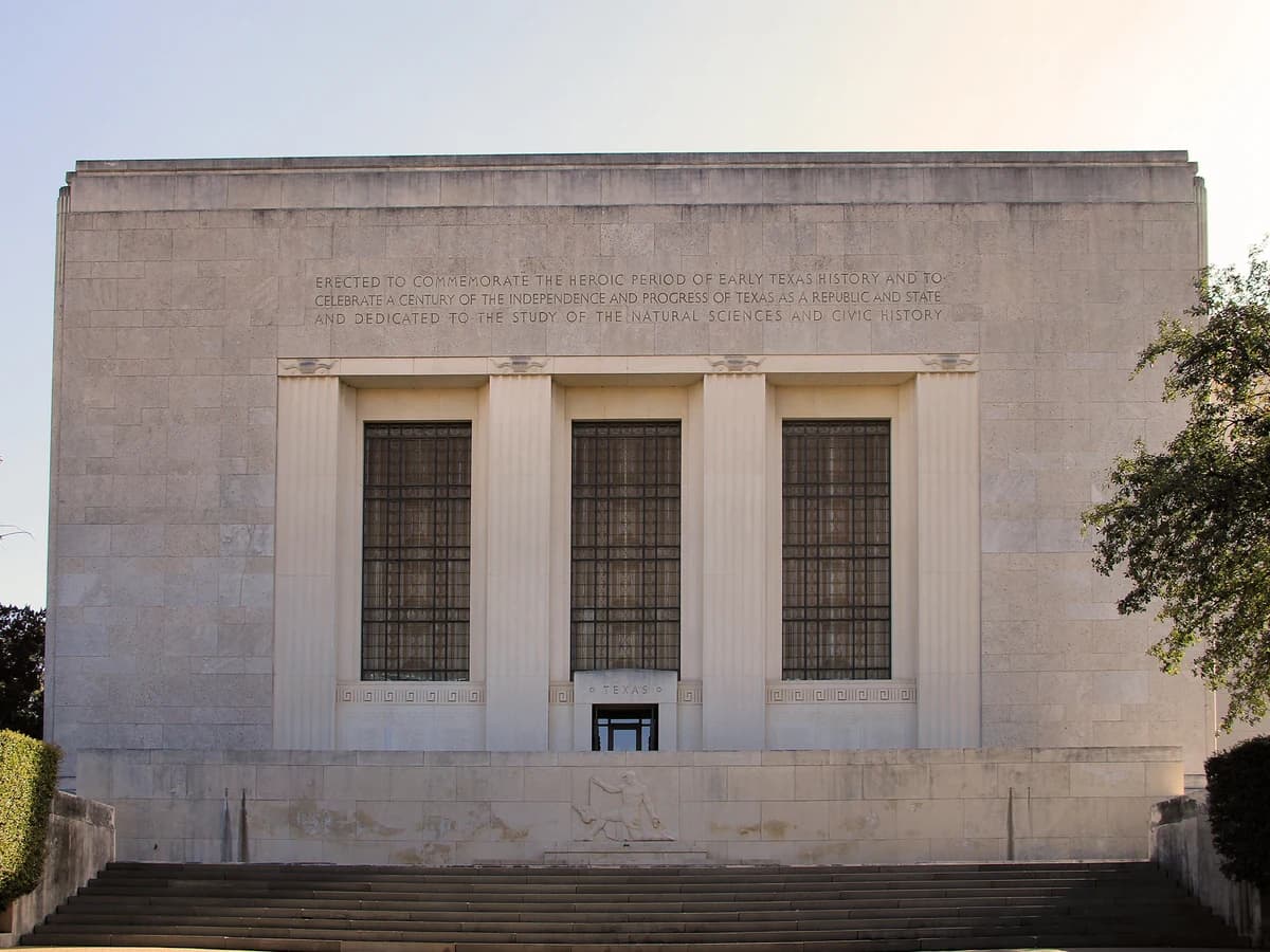 The pale limestone front of a museum with tall pilasters framing three barred windows under a carved inscription, above a wide flight of stone steps