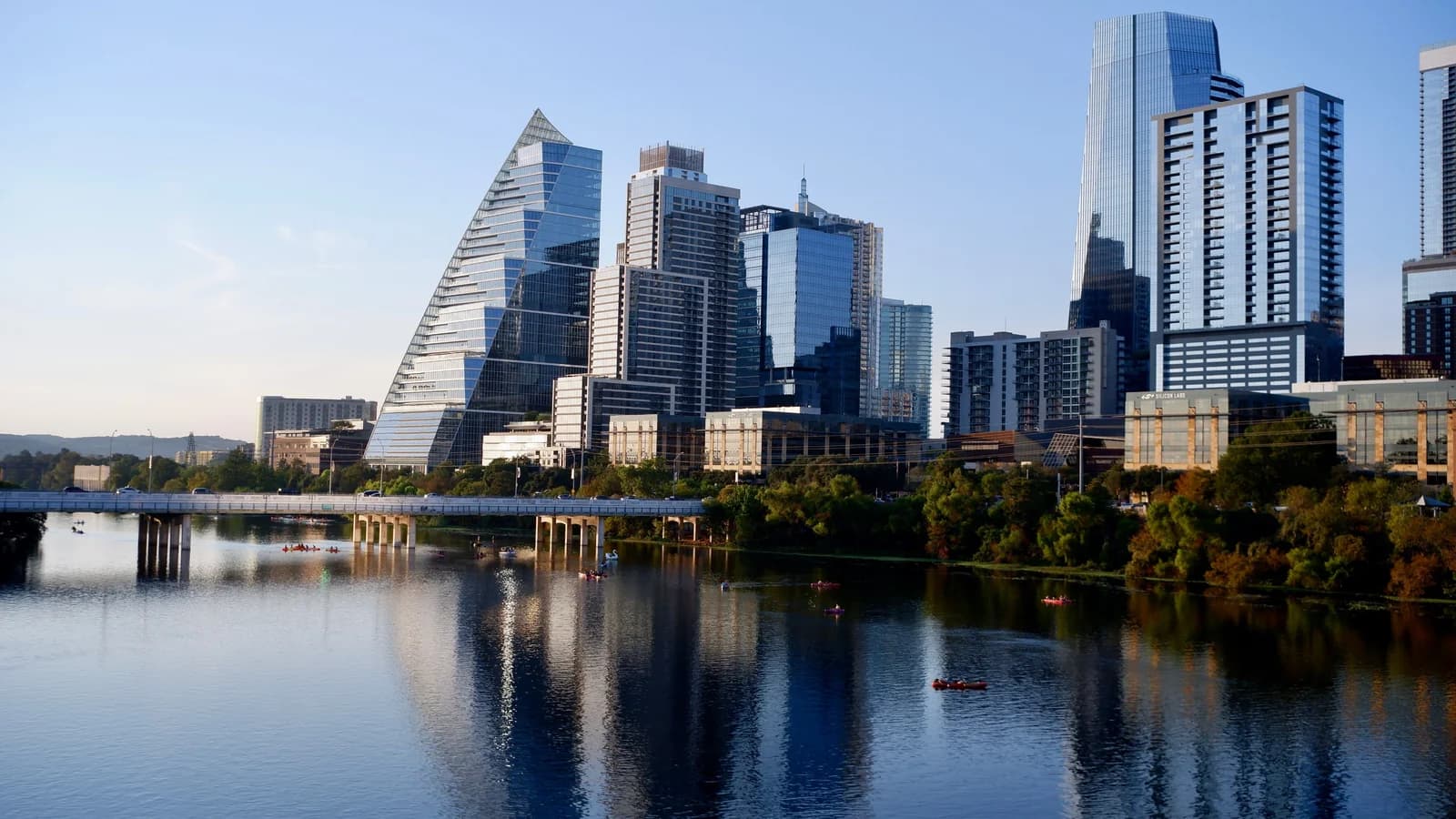 Downtown Austin towers, including a sail-shaped glass skyscraper, reflected in calm Lady Bird Lake with a bridge, kayakers and trees along the shore