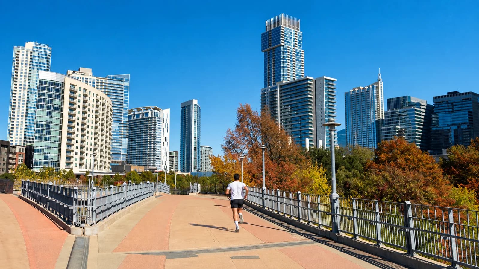 A runner in a white shirt crosses the Pfluger Pedestrian Bridge toward downtown Austin towers and autumn trees under a clear blue sky