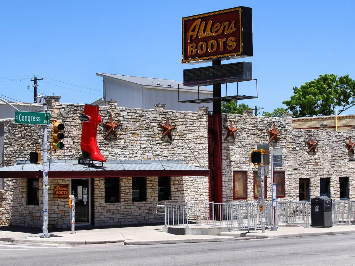 A long low stone storefront with a tall Allens Boots sign and a giant red cowboy boot on the corner, under a clear blue sky