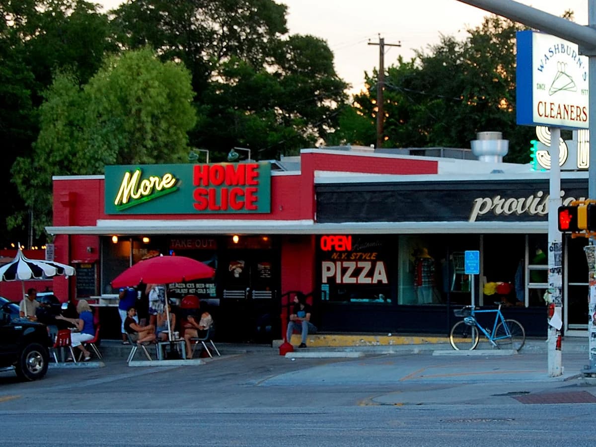 A red and white pizzeria storefront with Home Slice and Pizza signs, umbrellas over sidewalk tables and parked cars along the street