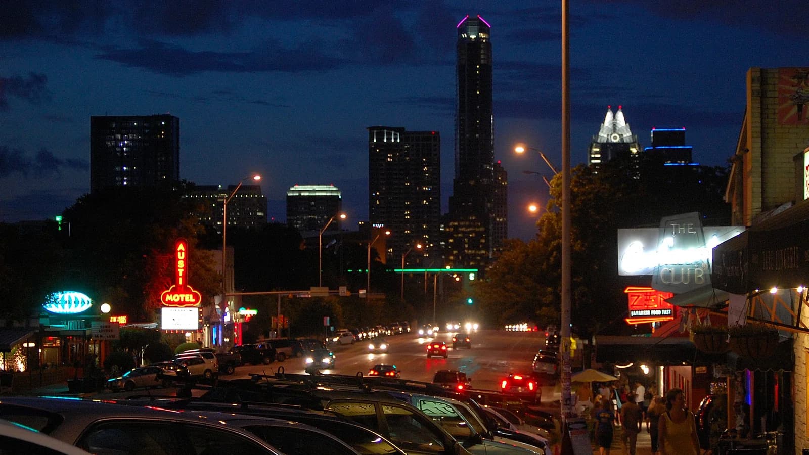 South Congress Avenue at night, with glowing neon motel and shop signs, parked cars along the street and downtown towers lit against a deep blue sky