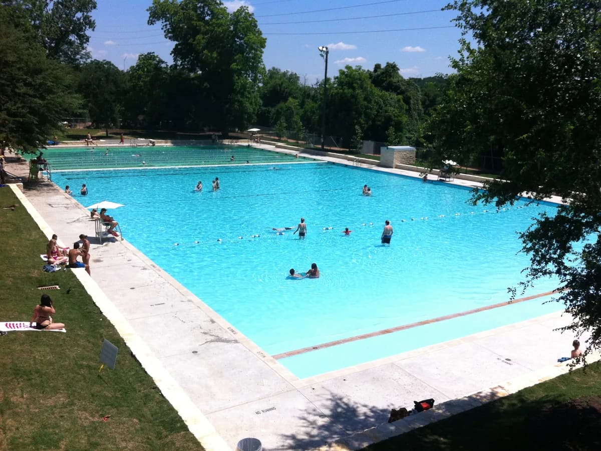 A long bright blue swimming pool with a wide concrete deck, a few swimmers and sunbathers on the grass, ringed by tall trees