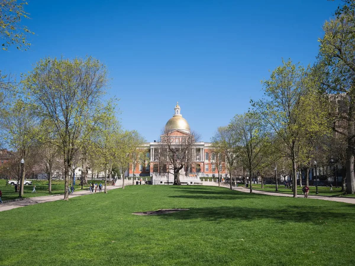 Wide green lawn of Boston Common with paths and spring trees leading to the gold dome of the Massachusetts State House under a clear blue sky