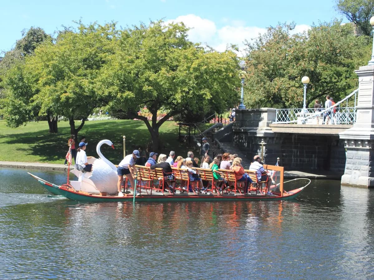 A swan boat full of riders gliding across the Public Garden lagoon toward the stone footbridge, with leafy trees along the grassy shore