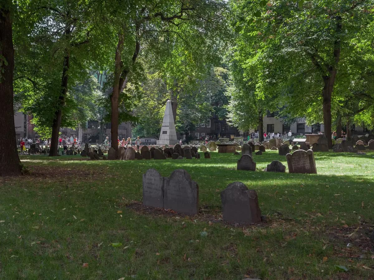 Old slate headstones on the shaded lawn of the Granary Burying Ground, with the granite Franklin obelisk among the trees behind