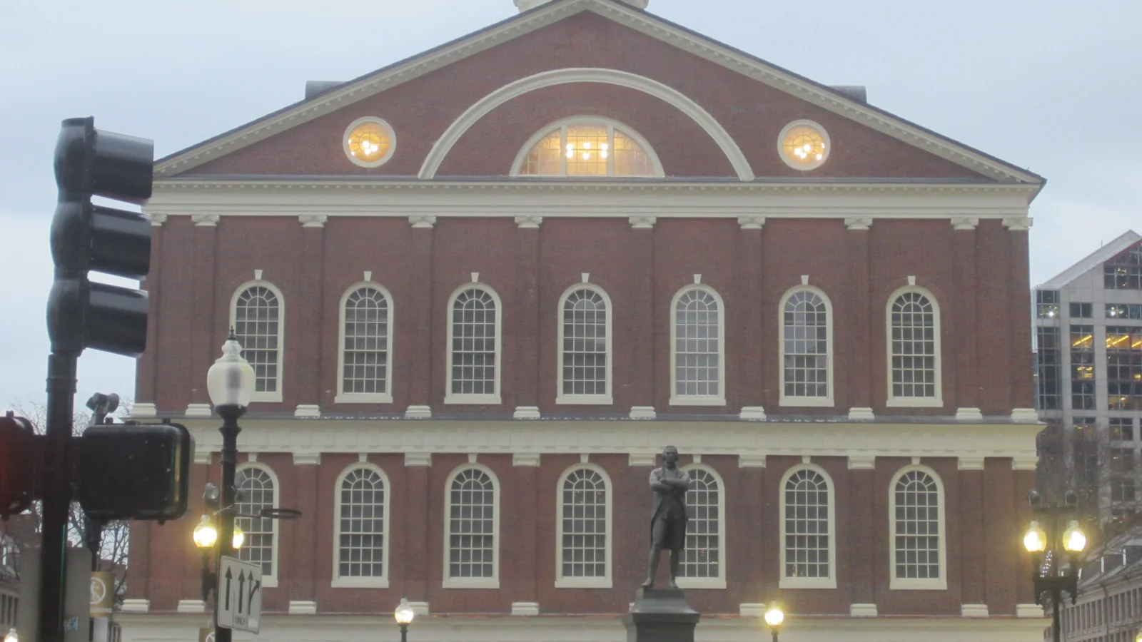 Red brick front of Faneuil Hall with its arched windows, lit lunettes and white cupola, with the Samuel Adams statue in front at dusk