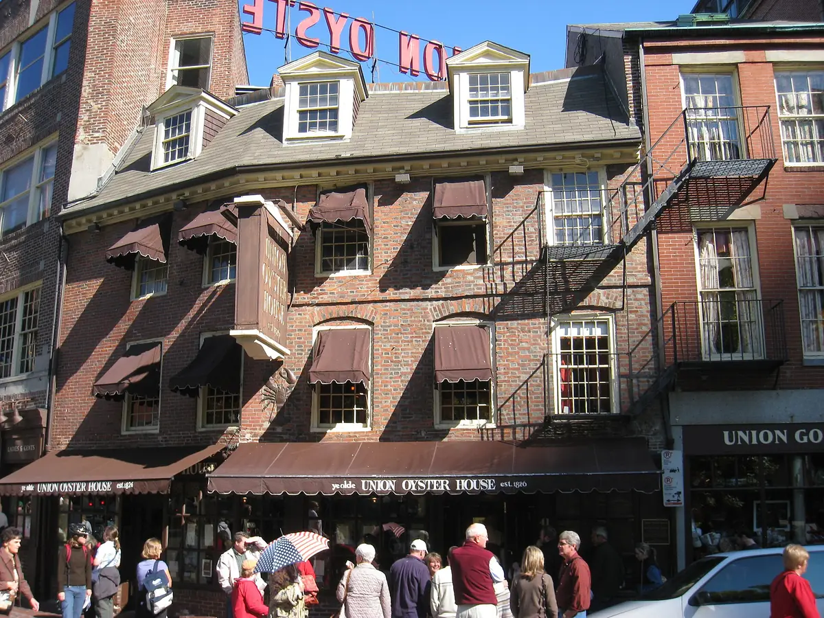 Old brick Union Oyster House with dormer windows, brown awnings and a red rooftop sign, with visitors gathered on the sidewalk out front