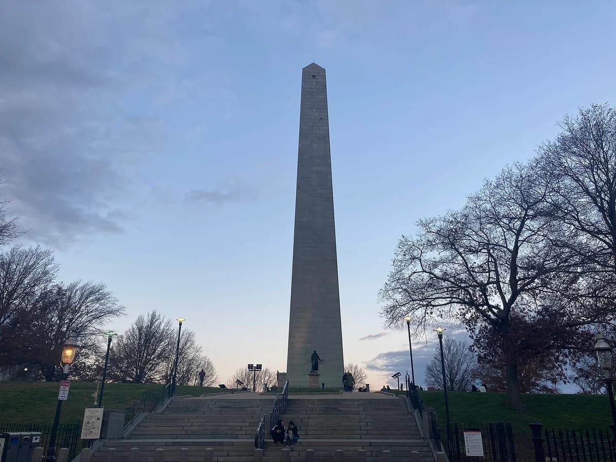 Granite obelisk of the Bunker Hill Monument rising from the hilltop at dusk, with a bronze statue at its base and wide stone steps in front