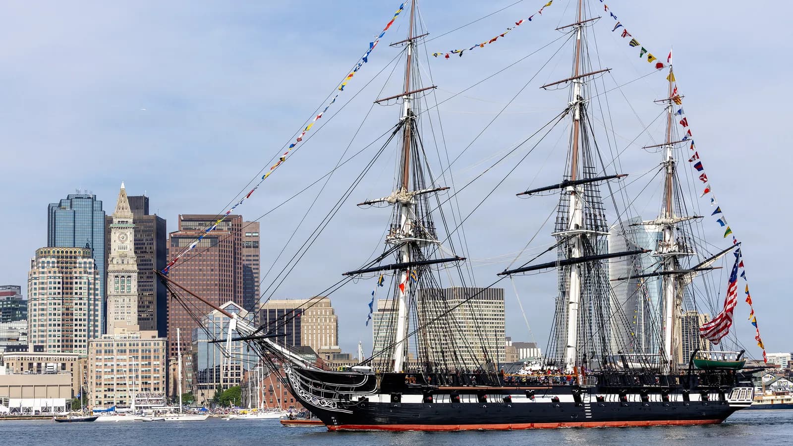 USS Constitution dressed in signal flags on Boston Harbor with the Custom House Tower and downtown skyline behind