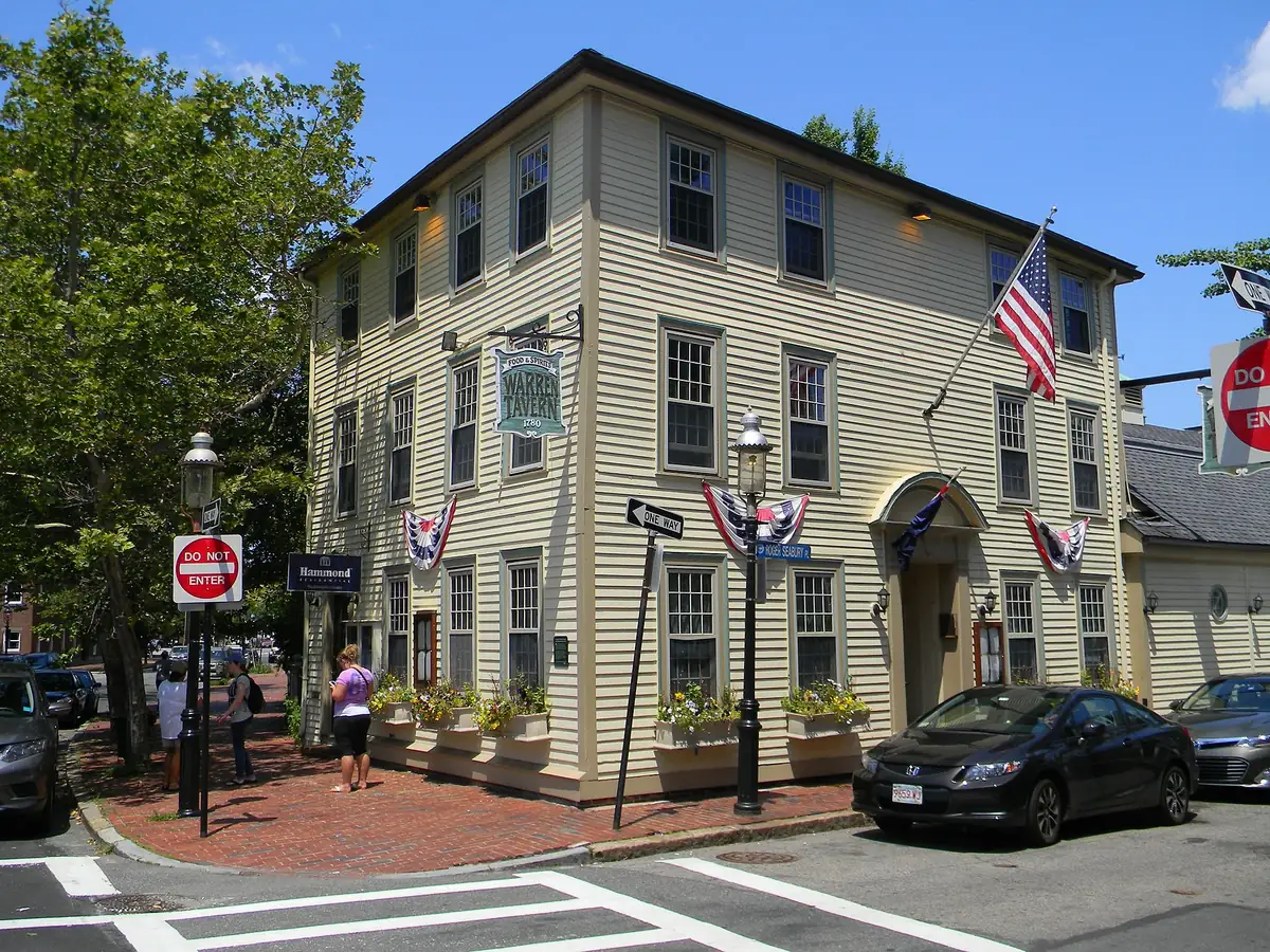 Pale yellow clapboard Warren Tavern on a Charlestown corner, with its hanging sign, an American flag, bunting over the windows and flower boxes along the brick sidewalk