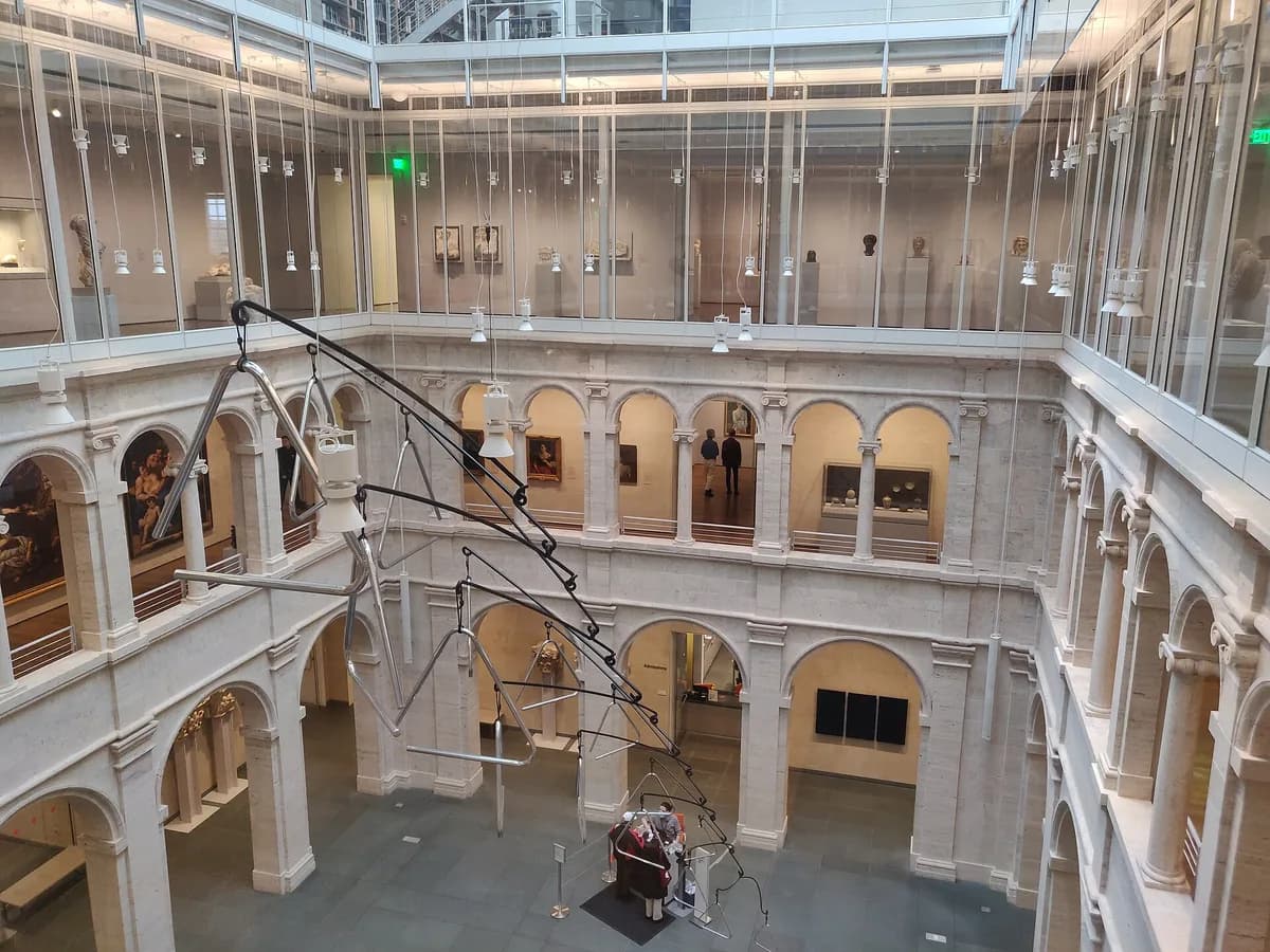 Pale stone arcaded courtyard of the Harvard Art Museums seen from an upper floor, ringed by glass walled galleries, with a hanging metal sculpture