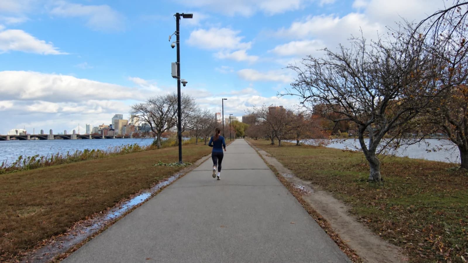 Woman in a navy top running the Charles River Esplanade path past bare fall trees, with the Longfellow Bridge and Boston skyline across the river
