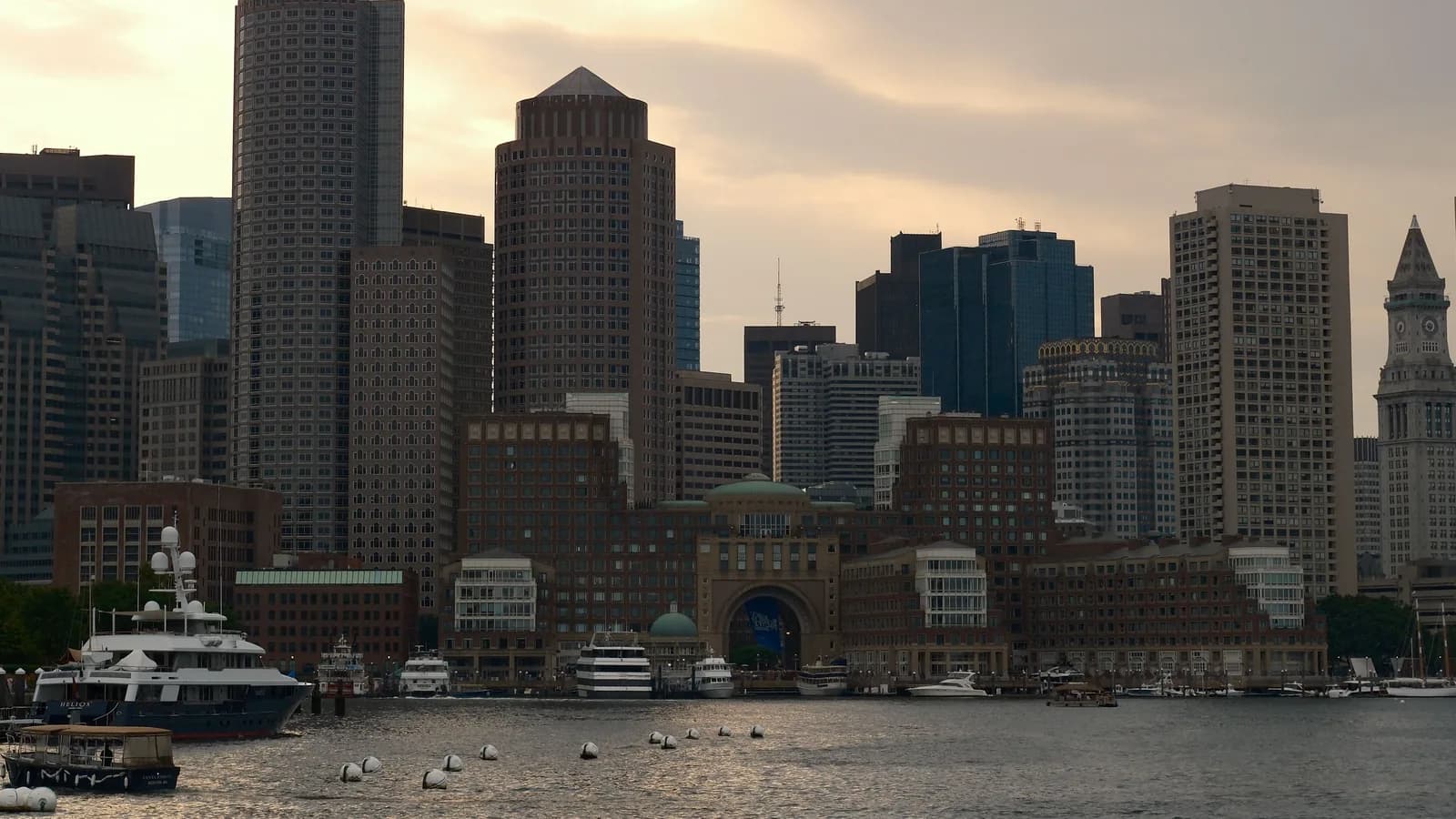 Boston skyline and the Rowes Wharf arch across the harbor at sunset, with boats moored along the waterfront